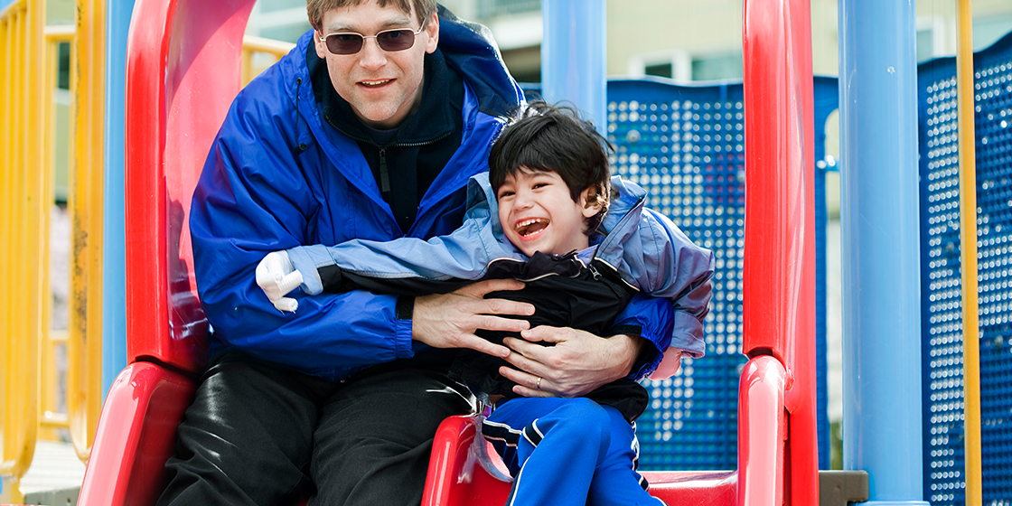 Father and young son on a playground slide. Son has cerebral palsy.