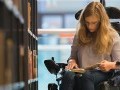 Teenage girl seated in a wheelchair, reading a book in a library.